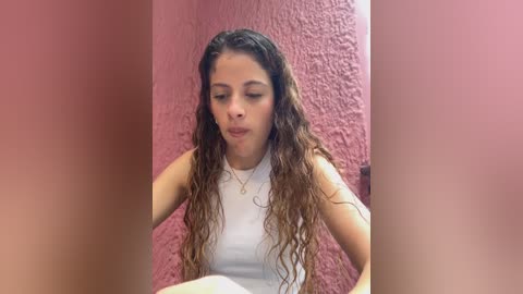 Media: A video of a young woman with curly, shoulder-length hair, wearing a white sleeveless top, sitting in a room with a pink textured wall. Her expression is neutral.