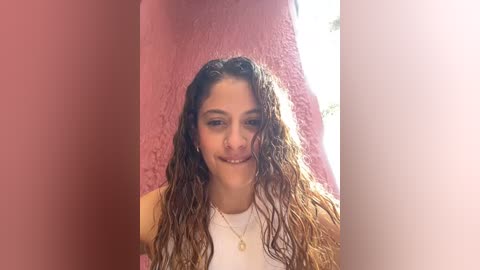 Media: Video of a young woman with long, wet, curly brown hair, light skin, wearing a white tank top, standing against a pink wall with sunlight filtering in from the right.