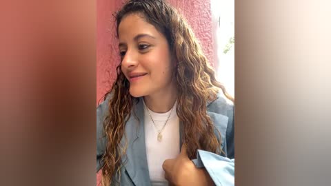 Media: Video of a young woman with curly brown hair, wearing a light blue blazer over a white shirt, standing against a pink wall, smiling softly.