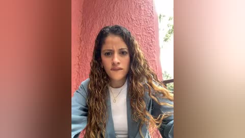 Media: Video of a young Latina woman with long, wavy, brown hair and a medium complexion, wearing a white T-shirt and light blue jacket, standing against a red and beige textured wall, looking serious.