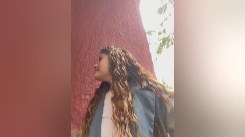 Media: Video of a young woman with long, wavy brown hair, wearing a blue jacket and white top, standing against a red and beige textured wall.