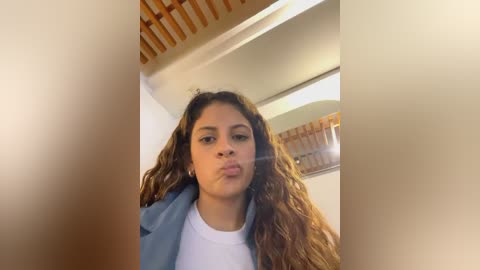 Media: Video of a young woman with long, wavy brown hair, puckering her lips, wearing a light blue jacket over a white shirt, indoors with wooden ceiling beams and a mirror in the background.