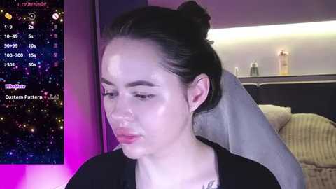 Media: A video of a young woman with pale skin and dark hair tied back, wearing a black top, sitting in a modern, dimly lit room with a starry background.