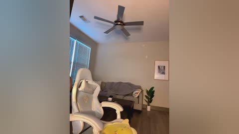 Media: Video of a modern, minimalist living room with beige walls, a gray sectional sofa, white gaming chair, and a ceiling fan.