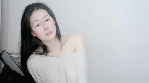 Media: A video of an East Asian woman with long black hair, fair skin, and a slender build, wearing a beige off-shoulder sweater, leaning against a plain white wall.