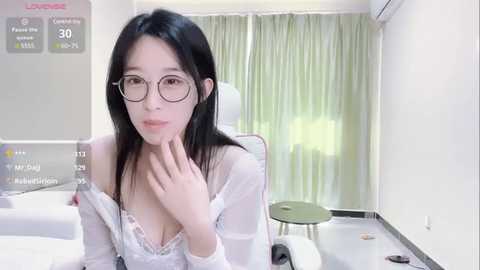 Media: Video of an Asian woman with fair skin, long black hair, and glasses, wearing a white lace bra, seated in a bright room with light green curtains.