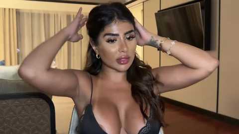 Media: Video of a curvy, light-skinned Latina woman with long, wavy hair, large breasts, wearing a black lace bra, adjusting her hair. She's in a modern, beige-toned room with a flat-screen TV on the wall and a bed in the background.