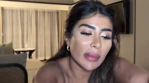 Media: Video of a woman with tan skin, dark hair, and bold black eyebrows. She's wearing makeup, sitting indoors with beige curtains in the background.