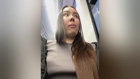 Media: Video of a young woman with long, straight brown hair and a light brown t-shirt, looking upward with a neutral expression, taken inside an airplane cabin.