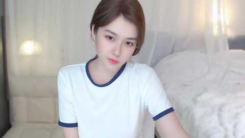 Media: Video of a young East Asian woman with fair skin, short brown hair, and red lipstick, wearing a white T-shirt with blue trim, sitting in a softly lit bedroom with white bedding and sheer curtains.