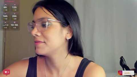 Media: A video of a light-skinned woman with straight black hair, wearing glasses, a black tank top, and a thin necklace, standing indoors. The background is a beige wall with a digital clock displaying 12:20.