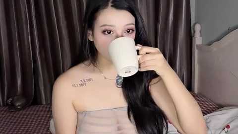 Media: Video of a young Asian woman with long black hair, wearing a sheer, strapless top, drinking from a white mug, with a tattoo reading \"I love you\" on her left shoulder, against dark curtains and a bed with red and white checkered bedding.