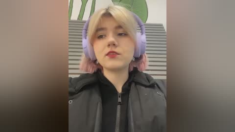 Media: Video of a young woman with pale skin, short blonde hair, and light purple headphones, wearing a black jacket. Background features a green leafy plant and a grey wall with horizontal stripes.