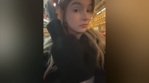 Media: A blurry video of a young woman with fair skin and brown hair, wearing a black top and brown coat, standing in a dimly lit, urban setting with blurred yellow and red lights in the background.