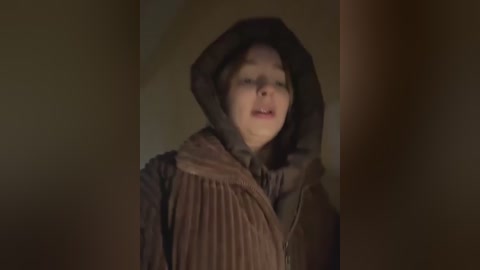 Media: Video of a young, fair-skinned girl with straight, shoulder-length brown hair, wearing a hooded brown cloak with a pleated tunic underneath, in a dimly lit, narrow hallway.