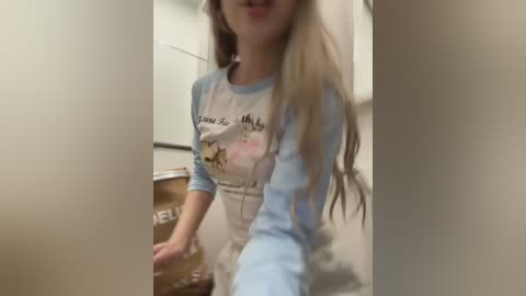 Media: Video of a young, fair-skinned woman with long, straight blonde hair, wearing a light blue and white graphic t-shirt with a cartoon character and apron, standing in a dimly lit, beige hallway.