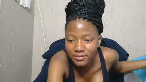 Media: Video of a young Black woman with braided hair in a dark blue tank top, leaning forward with a neutral expression against a beige wall.