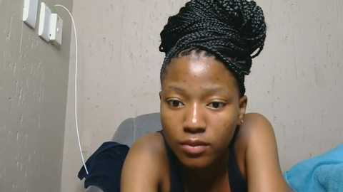 Media: Video of a young African woman with a braided hairstyle, wearing a black top, leaning forward, against a beige textured wall with electrical outlets.