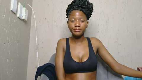 A video of a young Black woman with a slender physique and medium-sized breasts, wearing a black sports bra and a black head wrap, sitting against a textured beige wall with electrical outlets.