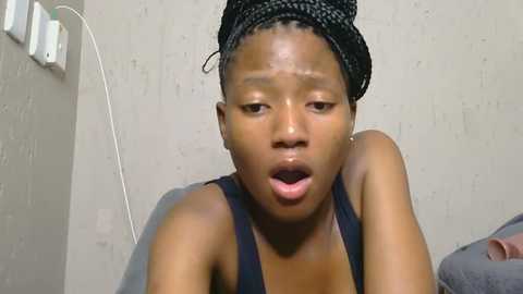 Video of a young Black woman with short, braided hair, wearing a black tank top, looking shocked, against a plain beige wall with an electrical outlet on the left.