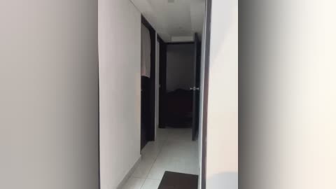 Media: A video of a narrow, dimly lit hallway with white walls, dark wooden doors, and a dark carpeted floor. The hallway leads to an open bedroom with a dark wooden bed and a white ceiling.