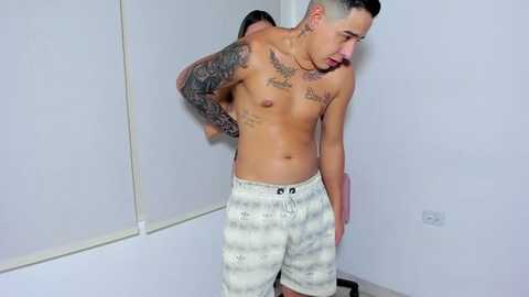 Media: Video of a tattooed, shirtless Latino man with short dark hair, wearing white patterned shorts, adjusting his waistband in a plain white room with mirrored walls.