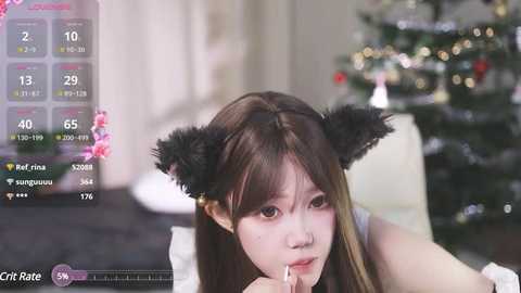 Media: Video of a young Asian woman with long brown hair and black cat ears, eating a lollipop indoors, next to a decorated Christmas tree.