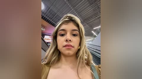 Media: Video of a young woman with light skin and long, blonde hair, wearing a beige top, standing in an indoor, modern setting with exposed metal beams and a blurred escalator in the background.