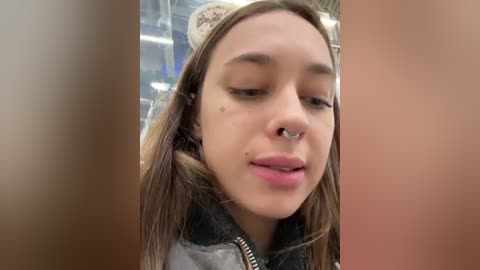 Media: Video of a young woman with light skin, brown hair, and a septum ring, wearing a gray jacket, looking slightly to the side. Background includes blurred, warm-toned walls.