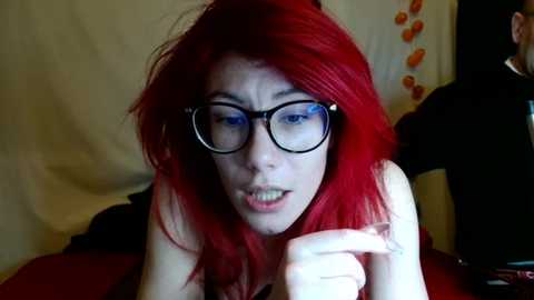 Media: Video of a young woman with bright red hair and glasses, lying on her stomach, looking at a phone, in a dimly lit room with a black wall and a white curtain with orange patterns.