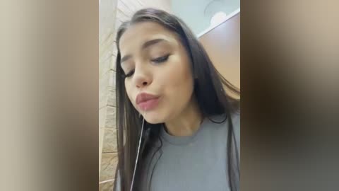 Media: Video of a young Latina woman with long dark hair, closed eyes, and pursed lips, wearing a light grey top, in a beige-tiled bathroom, looking introspective.