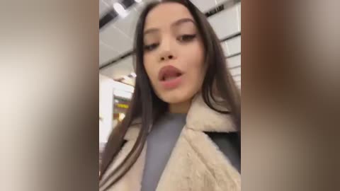 Media: Video of an Asian woman with long black hair, fair skin, and full lips, wearing a beige fur-trimmed coat over a light blue top, taken indoors.