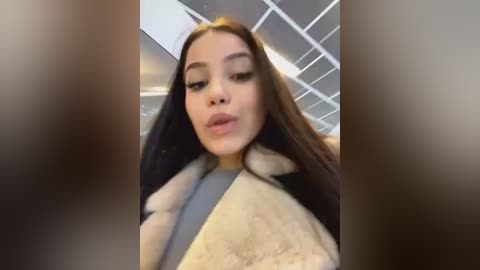 Media: A video of a young woman with long brown hair, fair skin, and full lips, wearing a fur-trimmed coat, taken indoors with a blurry background featuring a modern ceiling.