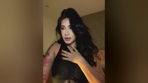 Media: Video of a young Latina woman with long black hair, wearing a black top, standing in a dimly lit room with beige walls. She has a tattoo on her left arm.