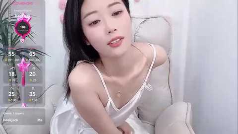 Media: Video of a young Asian woman with long black hair, fair skin, and a slender physique, wearing a white satin camisole, sitting on a white couch, with a digital health bar overlay.