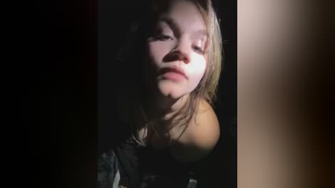 Media: Video of a young girl with light skin and shoulder-length, messy blonde hair, wearing a black top. She has a contemplative expression, partially obscured by shadows. The background is dark, creating a moody atmosphere.