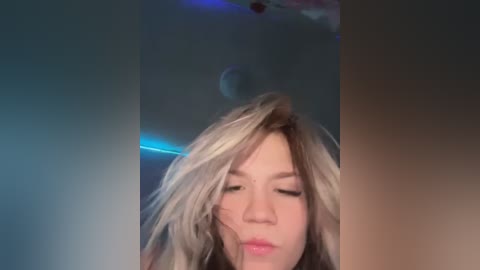 Media: Video of a young woman with blonde hair, wearing minimal makeup, in a dimly lit room with blue lighting. She has a neutral expression, with closed eyes and slightly parted lips.