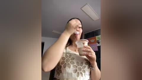 Media: A video of a woman with long brown hair, wearing a floral-patterned white dress, drinking a yogurt drink, in a dimly lit room with white walls and a ceiling light.