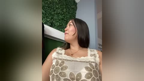 Media: Video of a woman with medium-length black hair, wearing a sleeveless, floral-patterned top, standing indoors near a green wall and white wall.