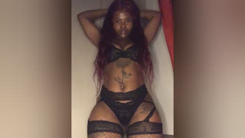 Media: Video of a dark-skinned woman with long, wavy red hair, wearing black lace lingerie, lying on a white bed, arms above head, in a dimly lit room.