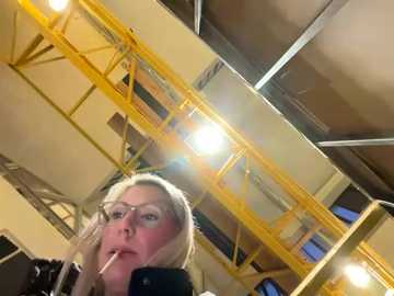 Media: A video of a blonde woman with glasses holding a cigarette, standing indoors in a warehouse-like setting with a yellow catwalk and industrial lighting.