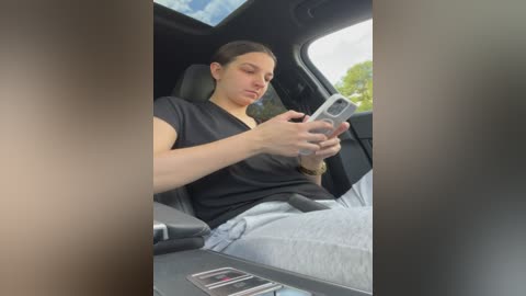 Media: A candid video captures a young woman with light skin and straight brown hair, wearing a black shirt and light blue jeans, seated in a car, engrossed in a smartphone.