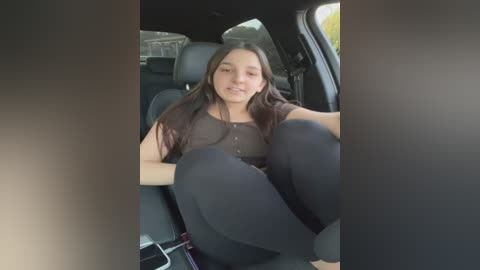 Media: Video of a young woman with long dark hair, seated in a car, wearing a black top and leggings, smiling softly, with a neutral background.