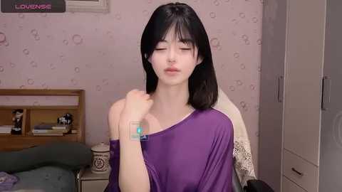 Media: Video of a young Asian woman with long black hair, wearing a purple off-shoulder top, holding a small blue box in her hand, in a softly lit bedroom with pastel wallpaper and a white cabinet.