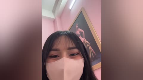 Media: Video of an Asian woman with long black hair, wearing a white face mask, in a pink room with a framed painting of a classical dancer.