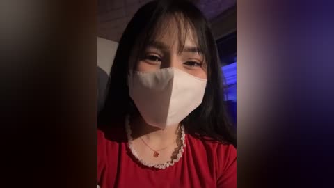 Media: Video of an Asian woman with long black hair, wearing a red blouse, a white surgical mask, and a beaded necklace, indoors with dim lighting.