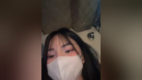 Media: A blurry video of a woman with long black hair and a white mask, lying on a pillow. The background is dimly lit, with indistinct objects.