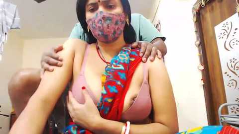 Media: Video of a young woman with medium skin tone and black hair, wearing a colorful saree and mask, receiving an injection from a man in a room with white walls and a wooden door.