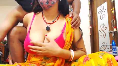 Media: Video of a topless South Asian woman with medium skin tone, wearing a bright pink bra, beaded necklace, and yellow saree, being held by a man in a dimly lit room.