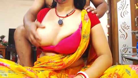 Media: Video of an Indian woman with medium skin tone, wearing a bright red blouse and yellow sari, her large breasts exposed, with a man's hands on her breasts. Background includes a TV, wooden furniture, and decorative items.